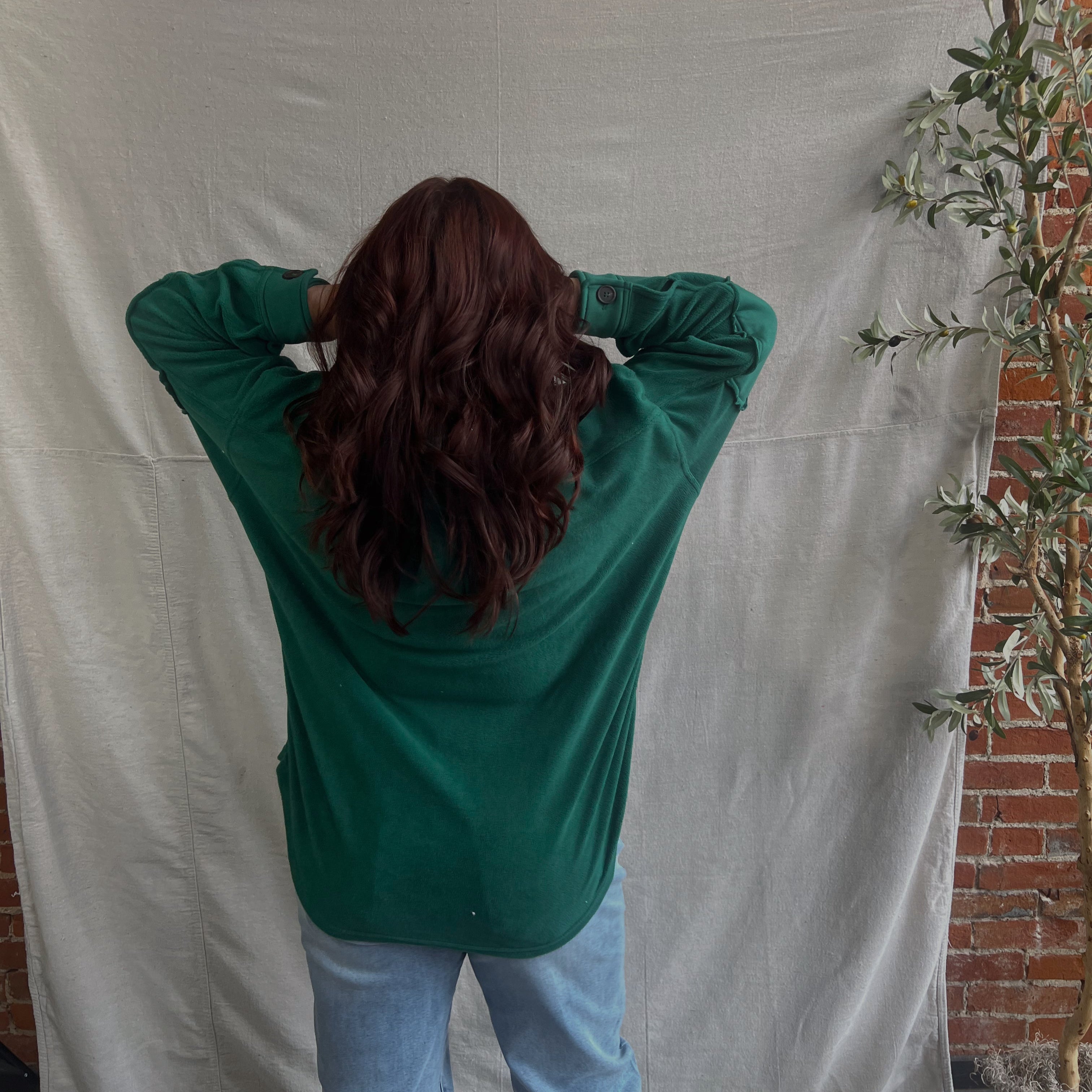 Person wearing a green shirt and light blue jeans standing against a white backdrop with a brick wall in the background.